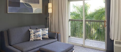 Quarto de hotel moderno com sofá cinza, um pequeno banquinho, abajur e uma janela grande com vista para palmeiras.