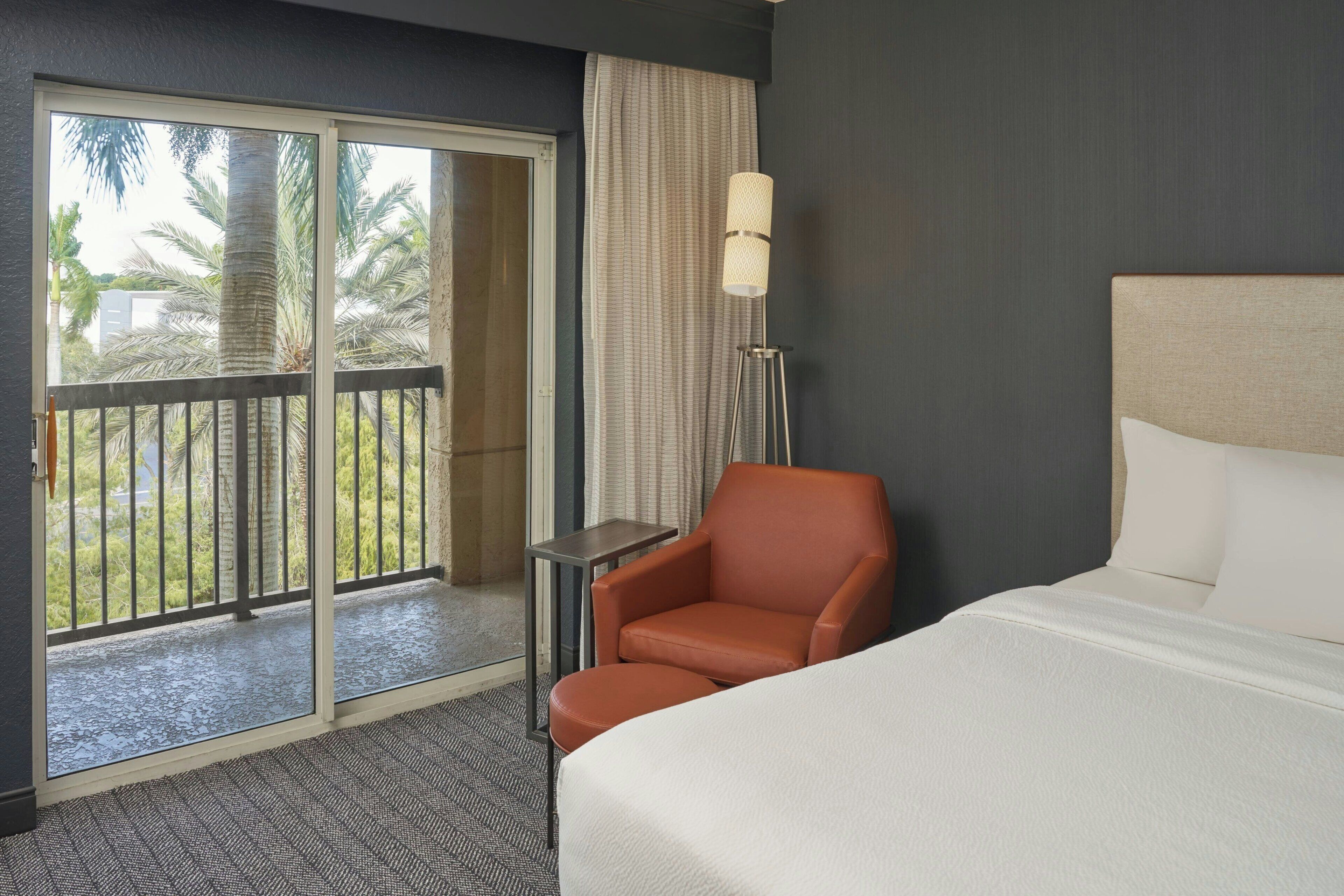 A hotel room with a bed, a chair, a lamp, a sliding glass door to a balcony, and a view of palm trees.