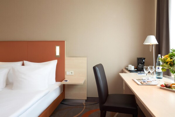 Une chambre d’hôtel avec un lit, un bureau, une chaise, une lampe, une machine à café, une bouteille d’eau, un vase avec des fleurs et une assiette avec de la nourriture.