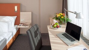 Une chambre d’hôtel avec un lit, un bureau avec un ordinateur portable, une chaise, une petite table avec un vase de fleurs et une fenêtre avec des rideaux.