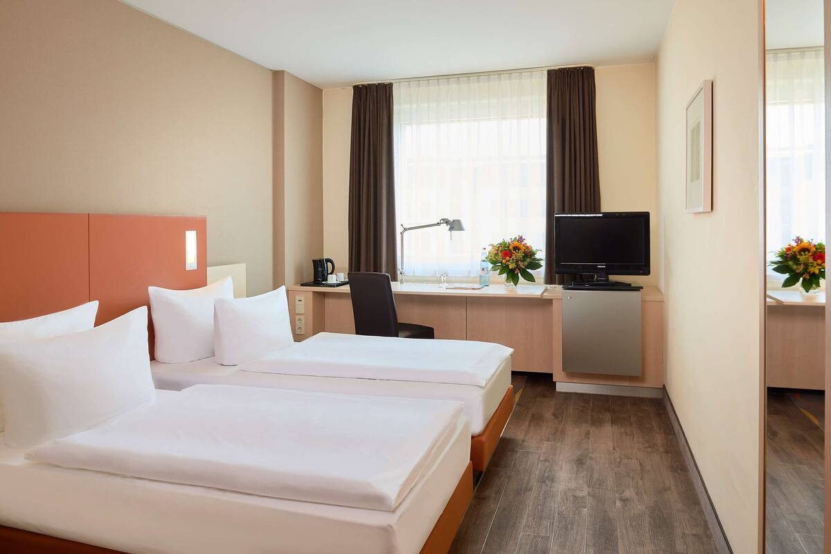 A hotel room with two beds, a desk with a computer and lamp, a TV, and a window with curtains.