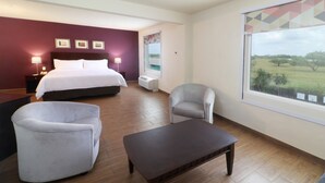 Une chambre d’hôtel comprenant un lit, deux fauteuils, une petite table, une table de chevet, une lampe, une fenêtre donnant sur un champ et un mur orné de trois tableaux.