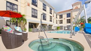 A swimming pool area with lounge chairs and an umbrella, surrounded by a building with multiple windows and a tiled roof.