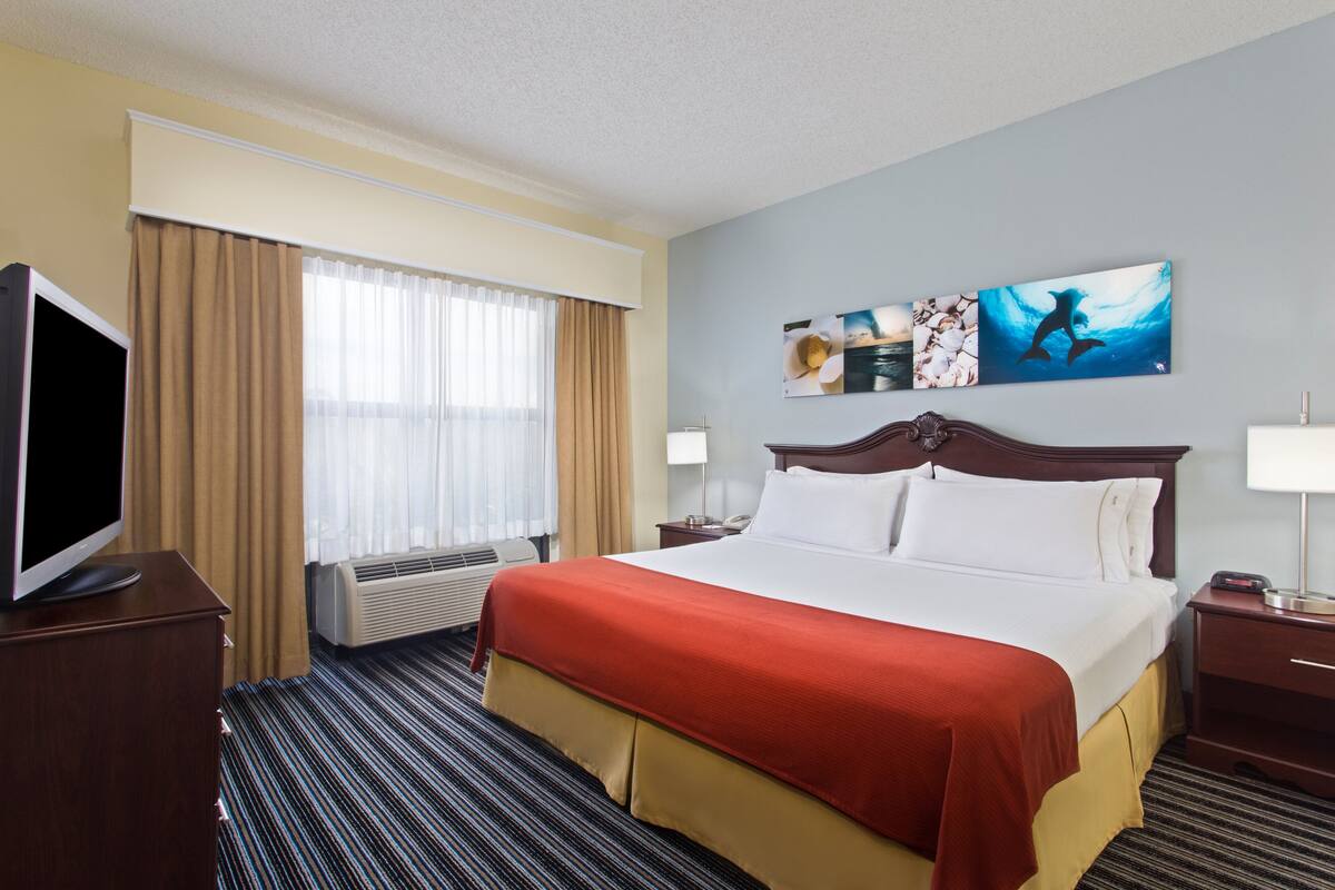 Ein Hotelzimmer mit einem großen Bett, einem Flachbildfernseher, einem Fenster mit Vorhängen und Bildern an der Wand.
