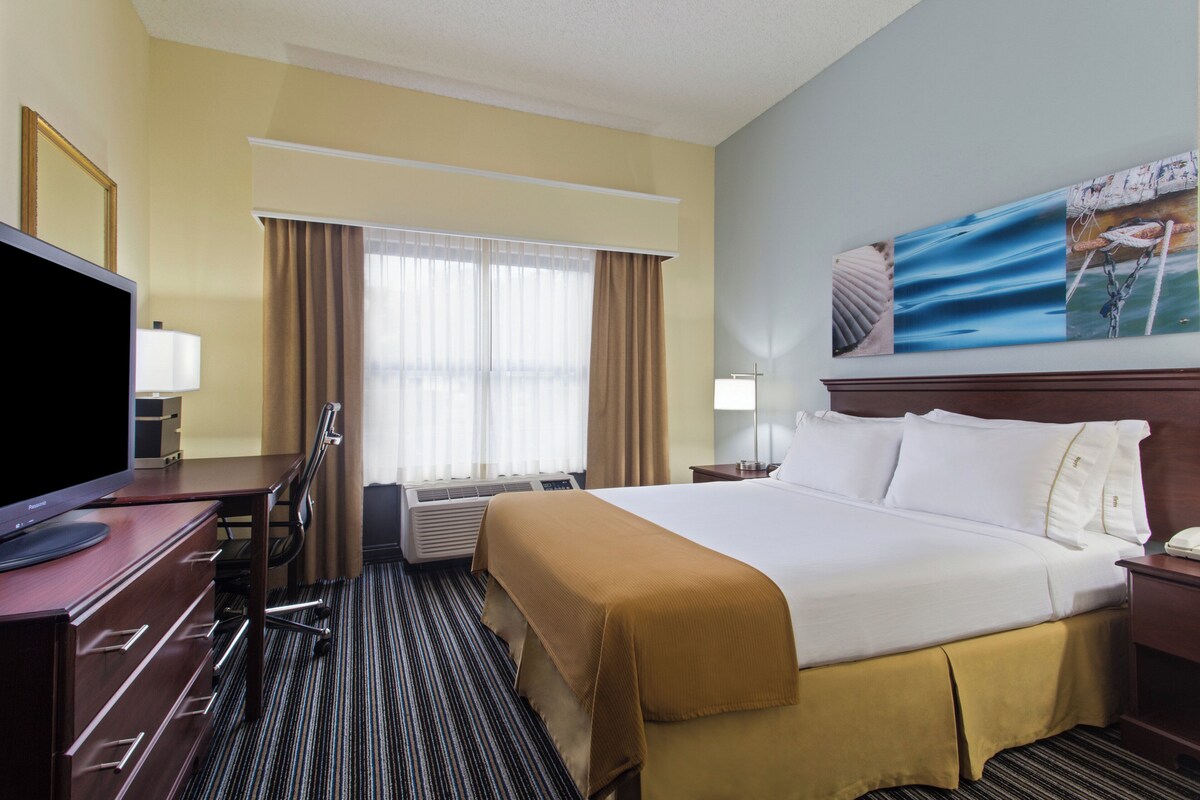 Ein Hotelzimmer mit einem Bett, einem Schreibtisch mit Computer, einem Fernseher, einem Fenster mit Vorhängen, einer Klimaanlage und einem Bild an der Wand.