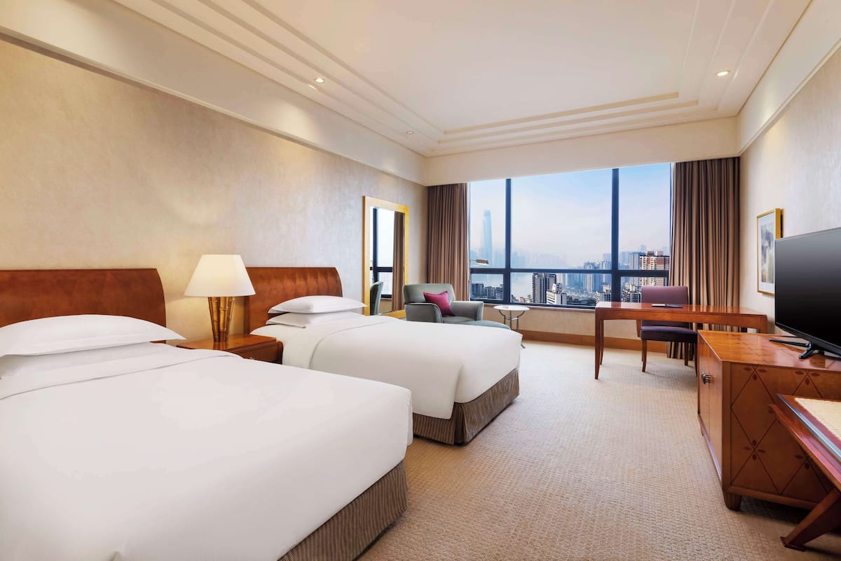 Een hotelkamer met twee bedden, een bureau, een televisie en een groot raam met uitzicht op de stad.
