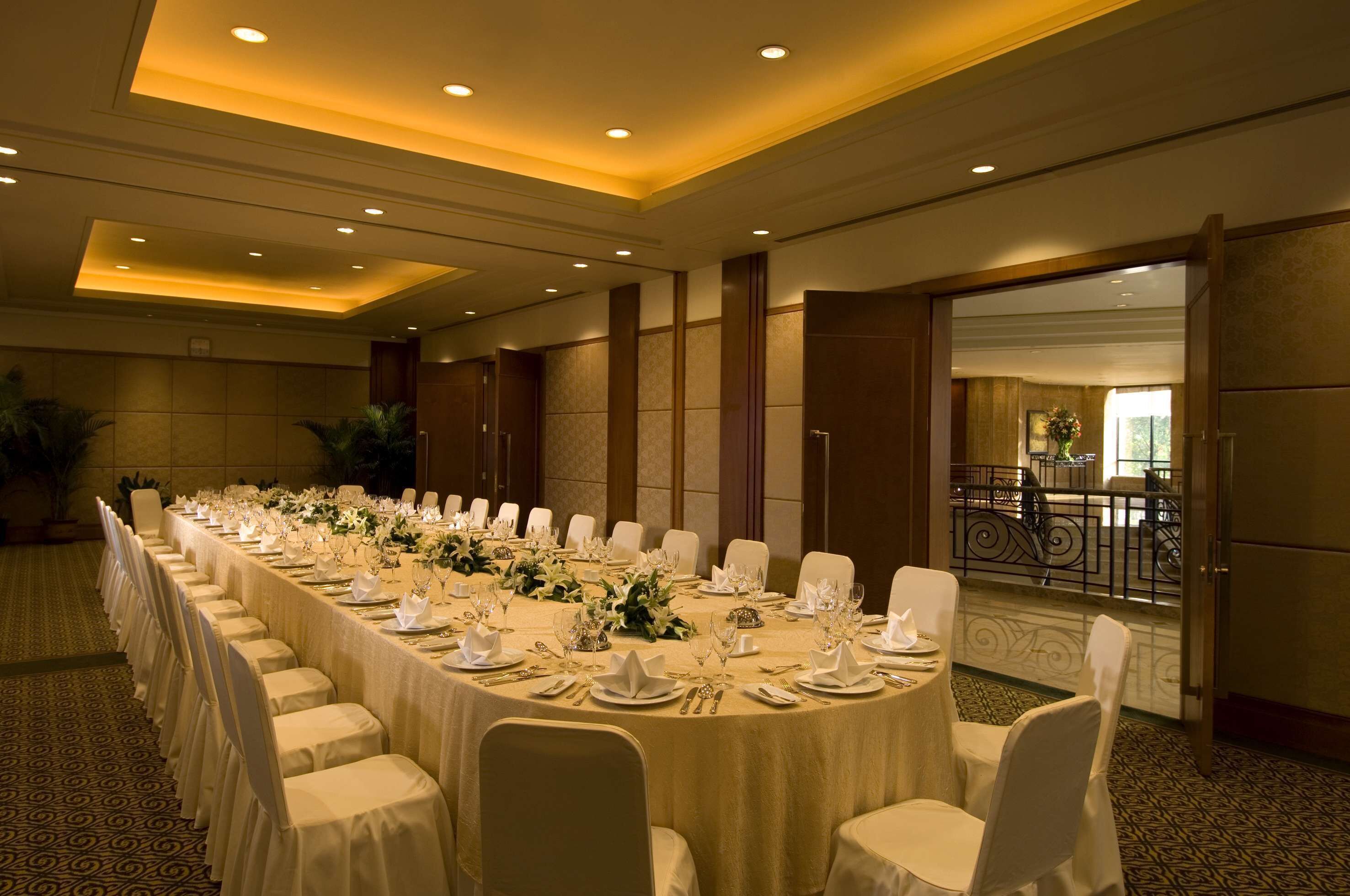 A long banquet table set with white tablecloths, chairs, and floral centerpieces in a well-lit room with a balcony view.