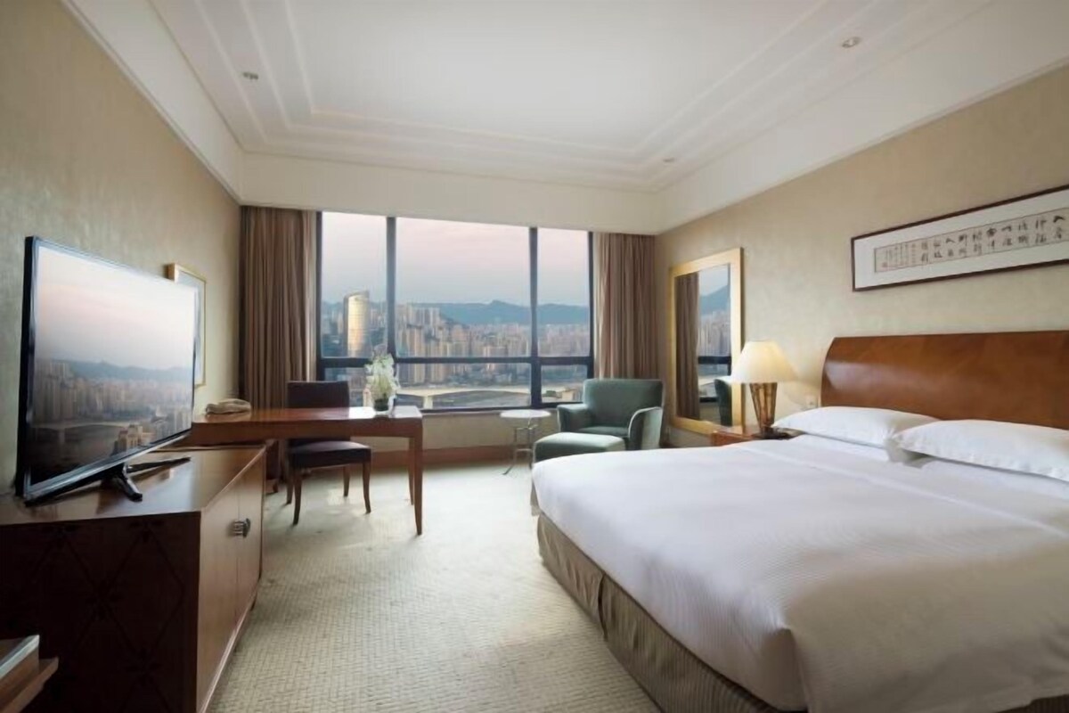 A hotel room with a large bed, a desk with a chair, a television, and a view of a cityscape through large windows.
