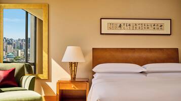 A hotel room with a bed, a nightstand with a lamp, a green armchair, a large window with a city view, and a framed calligraphy piece on the wall.