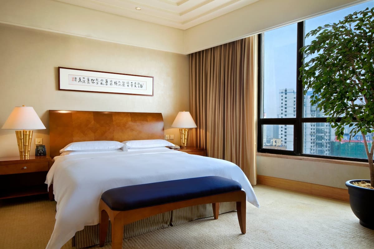 Een hotelkamer met een groot bed, twee bedlampen, een kunstwerk in een lijst aan de muur, een raam met gordijnen en een kamerplant.