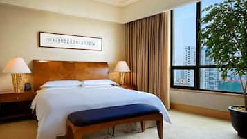 Habitación de hotel con una cama grande, dos lámparas de noche, una obra de arte enmarcada en la pared, una ventana con cortinas y una planta en maceta.