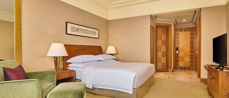 A hotel room with a bed, two bedside lamps, a green armchair, a wooden dresser, and a television.