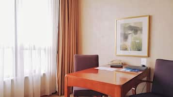 A hotel room with a wooden desk, two chairs, a framed picture on the wall, and a window with curtains.