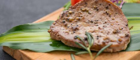 A steak with seasoning on a wooden board, garnished with a sprig of rosemary, and surrounded by vegetables.