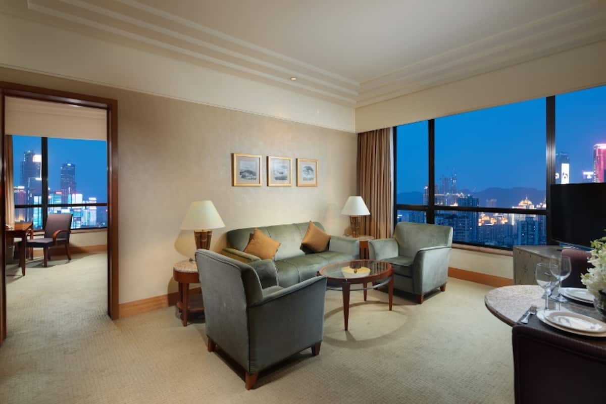 Hotelkamer met stadszicht, een bank, twee fauteuils, een salontafel, een eethoek met een tafel gedekt voor twee personen, en een televisie.