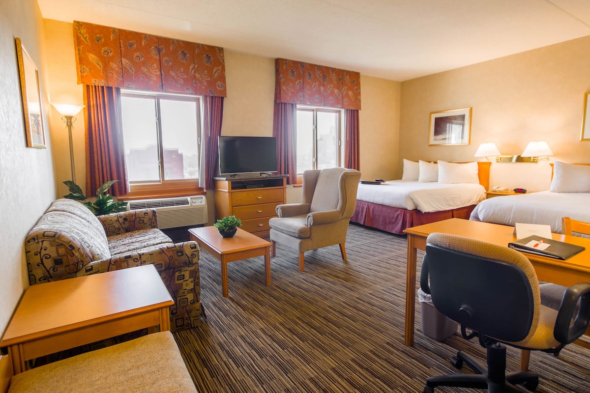 A hotel room with a sofa, armchair, desk, and a bed with two pillows.