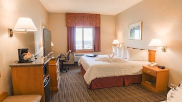 A hotel room with a bed, a desk with a computer, a chair, a TV, a window with curtains, a bathroom with a sink and mirror, and a painting on the wall.