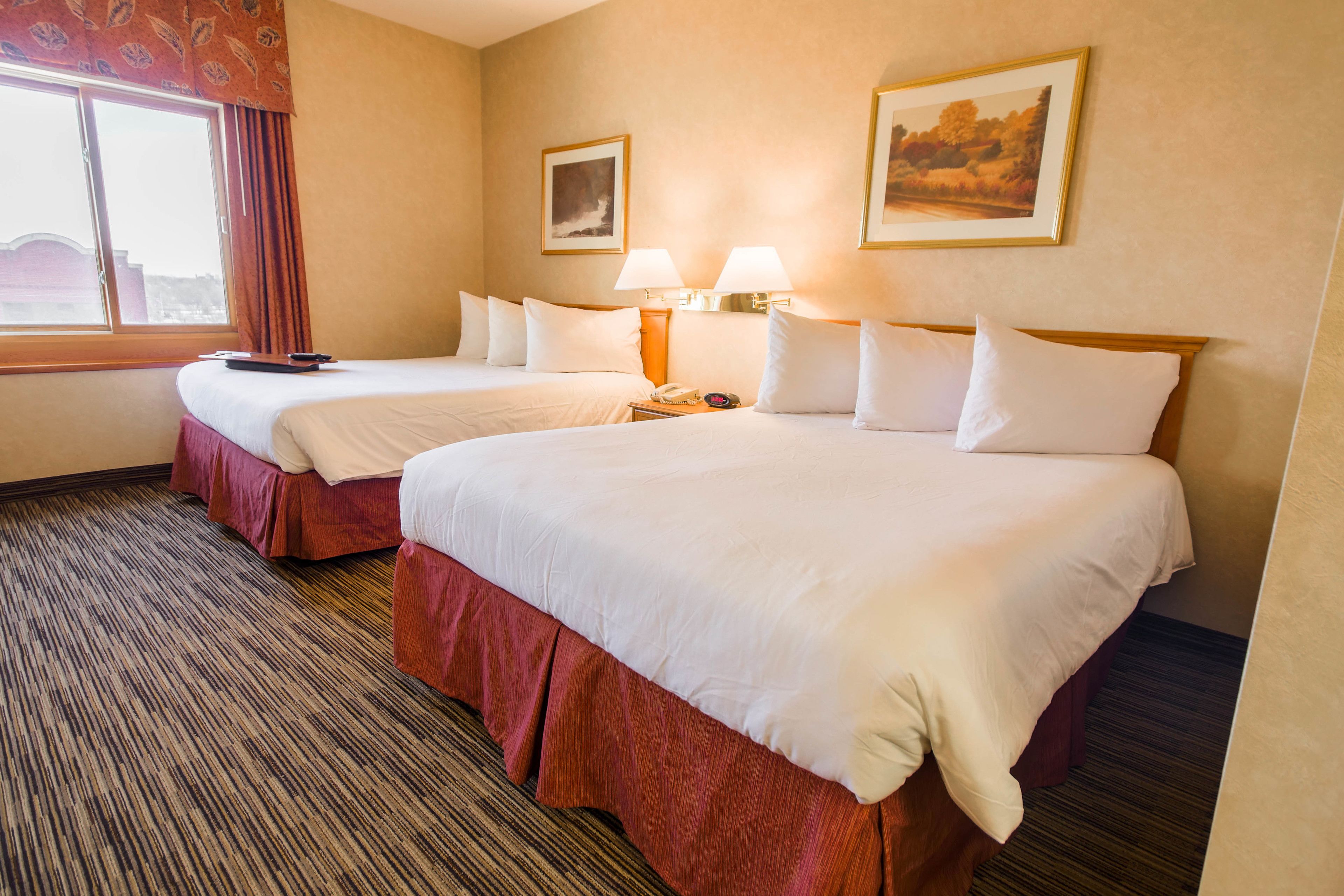 A hotel room with two beds, a carpeted floor, a window with curtains, and two wall-mounted lamps.