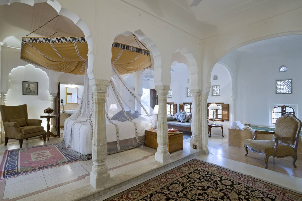 Um quarto espaçoso com uma cama grande, uma área de estar com sofá e poltronas, e vista através de uma porta arqueada.