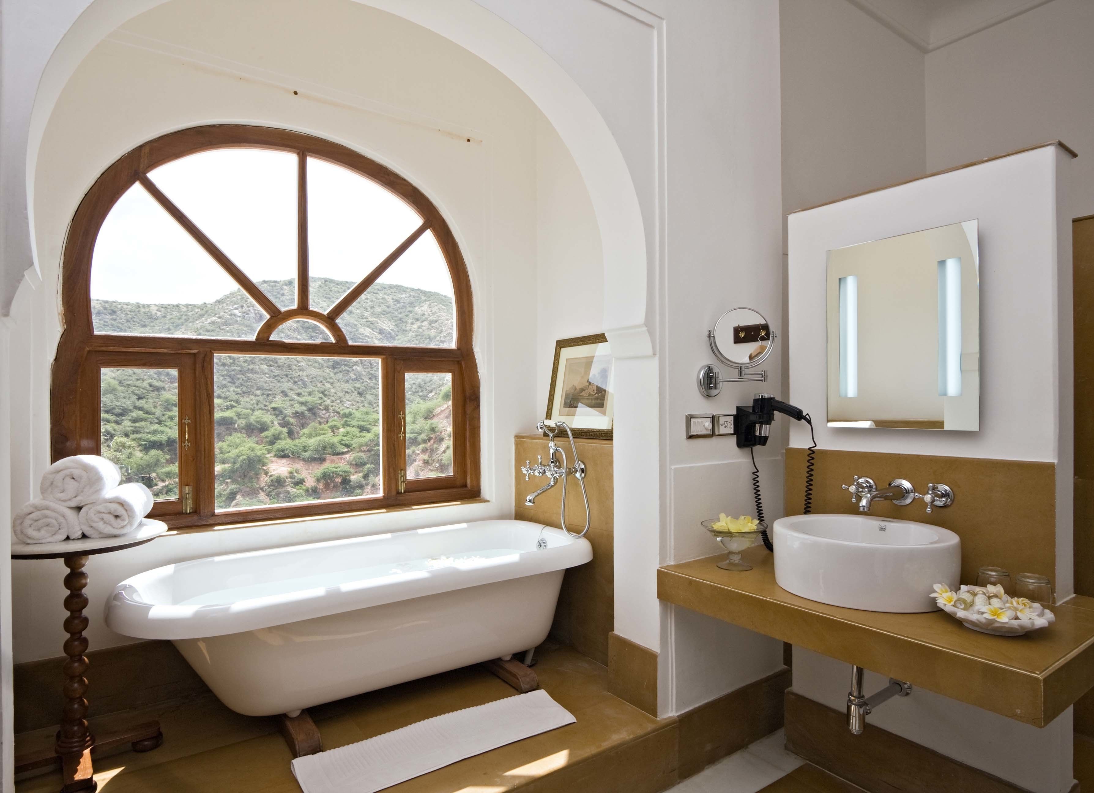 Une salle de bain avec une baignoire sur pieds, un lavabo et un grand miroir. Une fenêtre offre une vue sur la verdure à l’extérieur.
