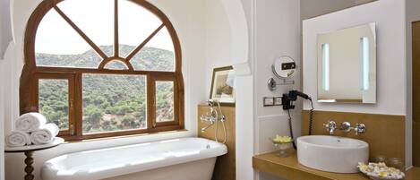 Une salle de bain avec une baignoire sur pieds, un lavabo et un grand miroir. Une fenêtre offre une vue sur la verdure à l’extérieur.