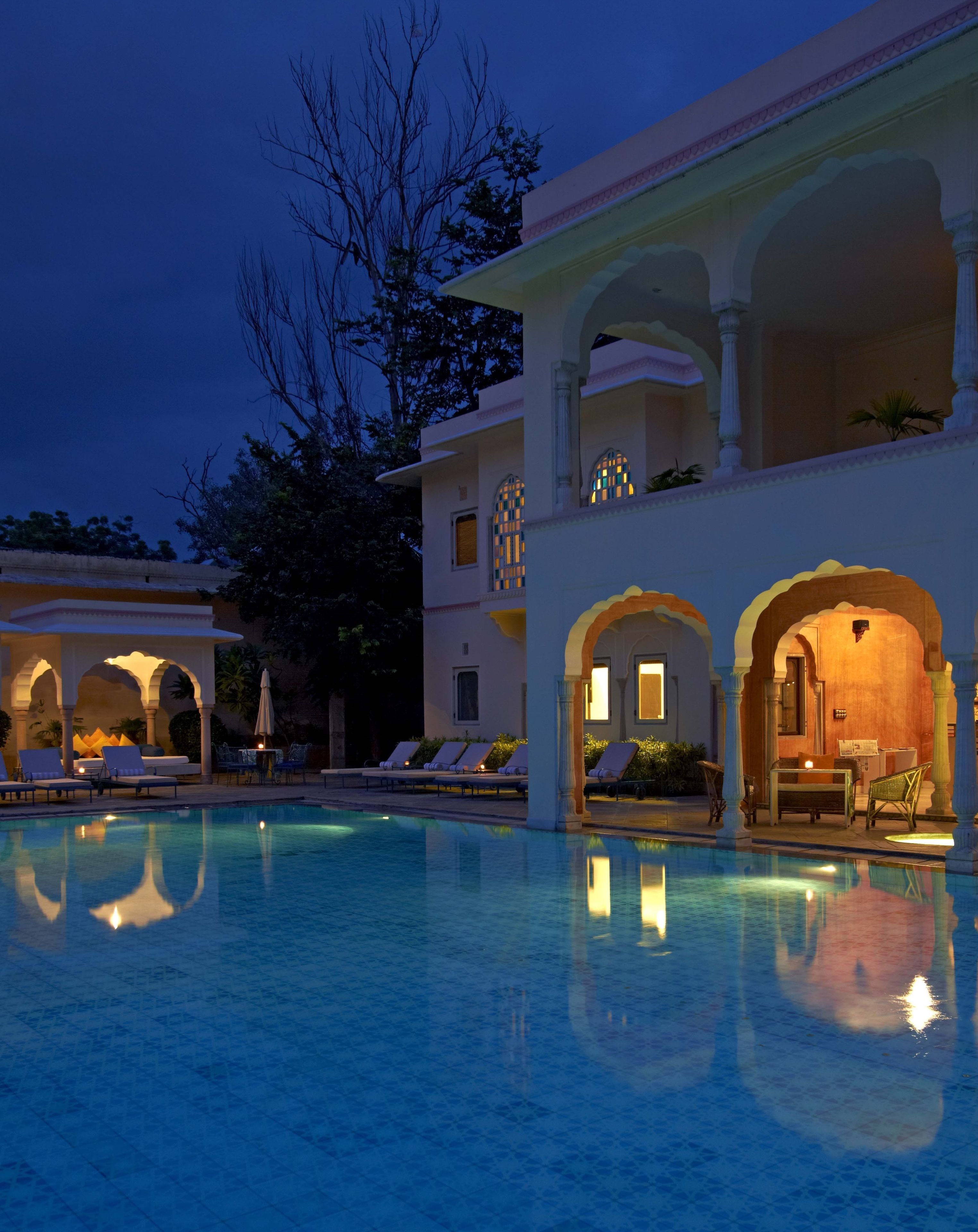 Zona de piscina con sillones reclinables y un edificio con ventanas arqueadas y columnas.