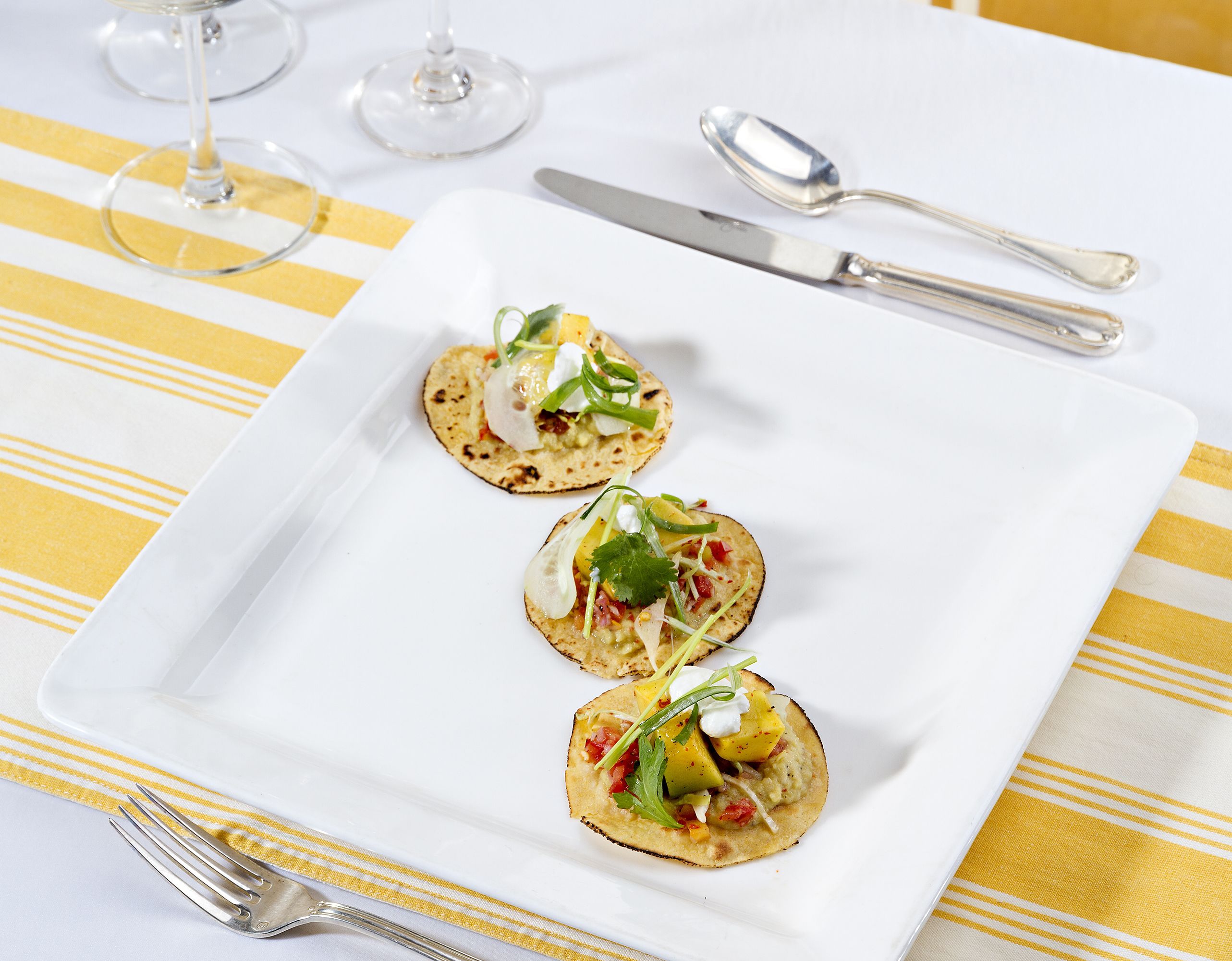 Un plato blanco cuadrado con tres aperitivos sobre una mesa con mantel a rayas amarillo y blanco.