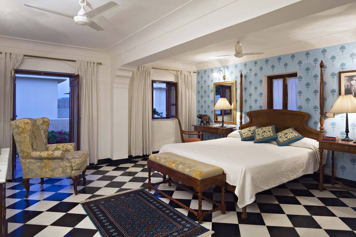 A hotel room with a checkered floor, a bed with a wooden headboard, a chair, a desk with a lamp, and a window with curtains.