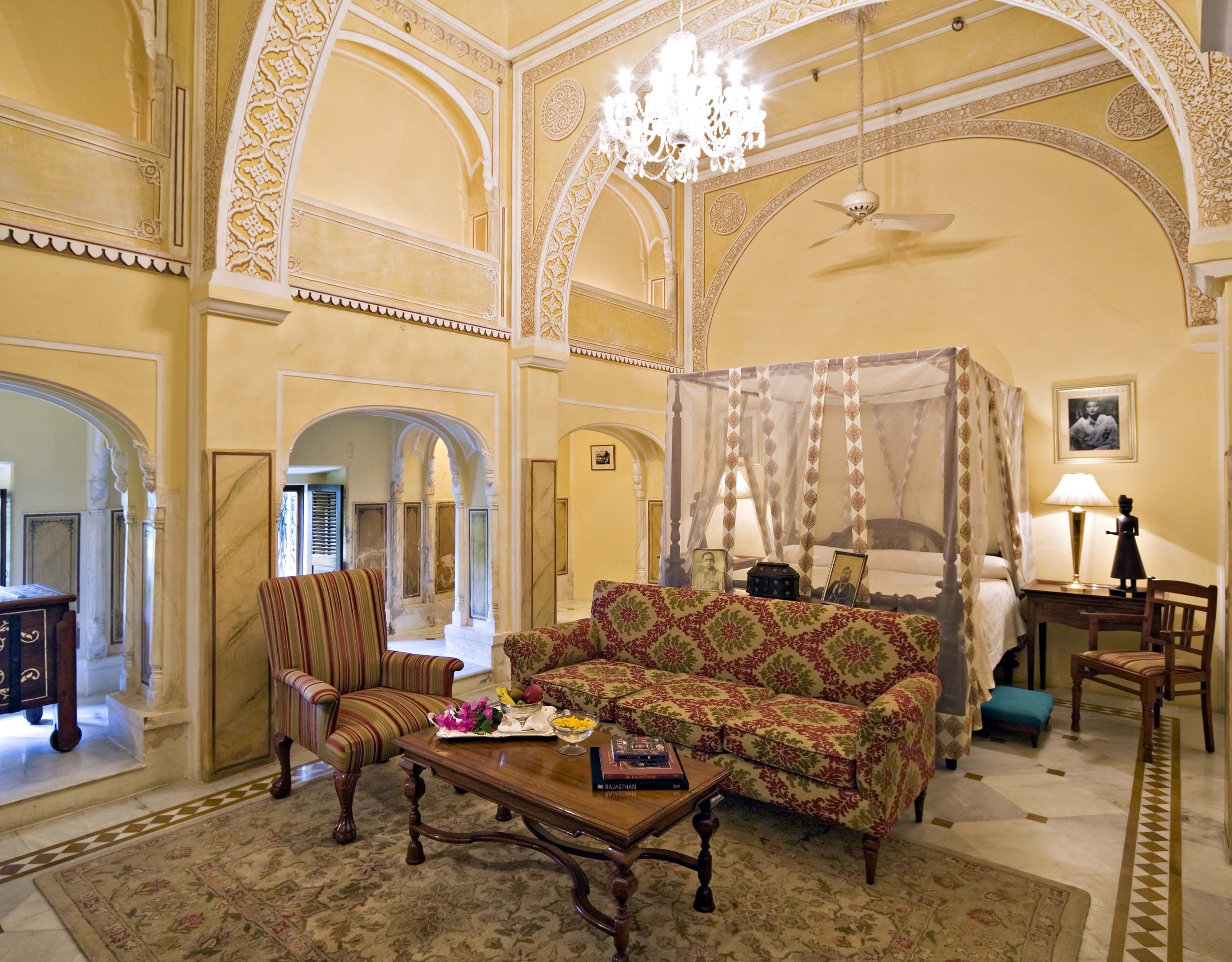A spacious room with a large sofa, a chair, a coffee table, and a canopy bed. There is a chandelier and a ceiling fan.