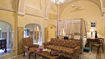 A spacious room with a large sofa, a chair, a coffee table, and a canopy bed. There is a chandelier and a ceiling fan.