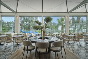 Un'ampia zona pranzo all'aperto con tavoli rotondi, sedie in vimini e un albero al centro.