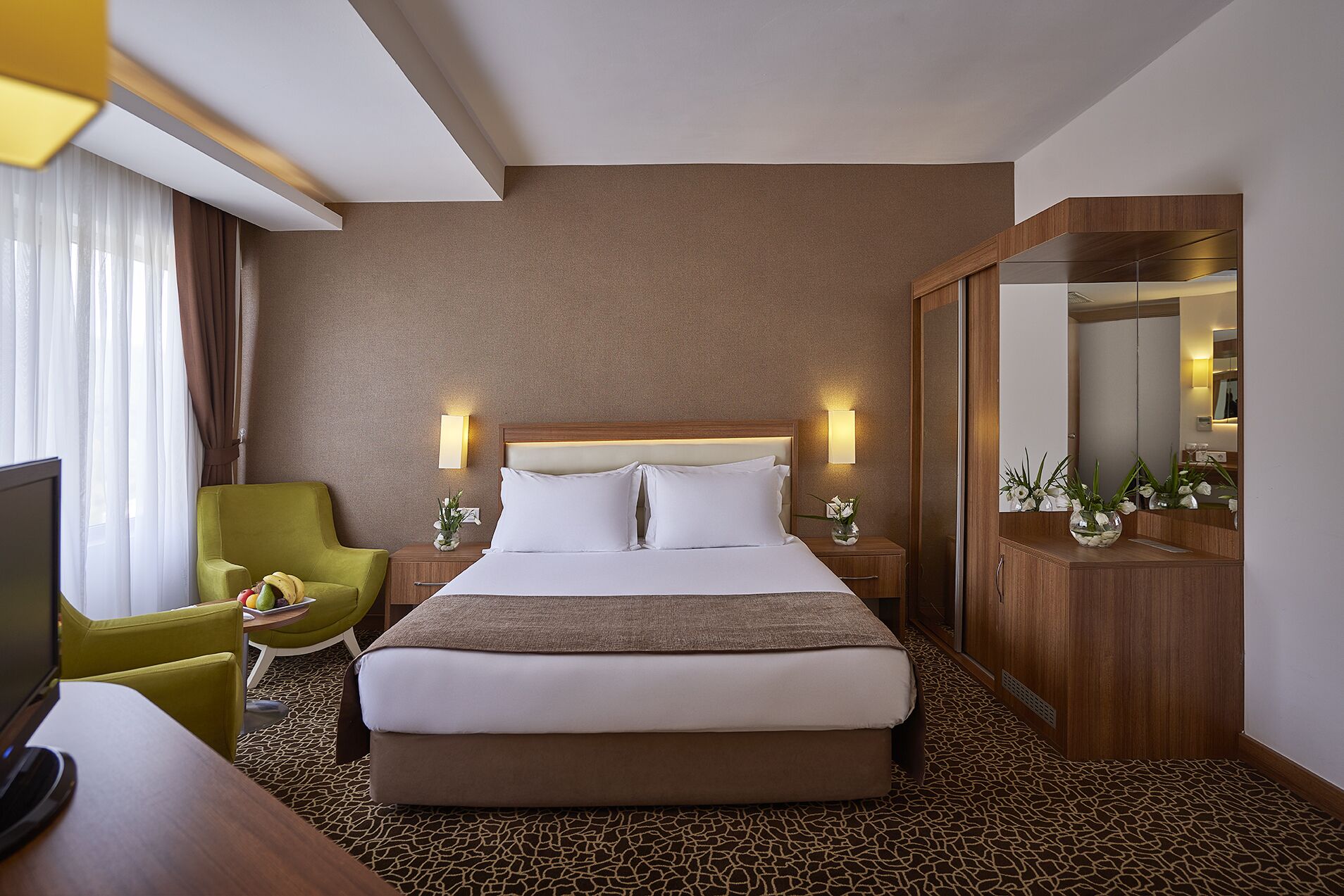 A hotel room with a large bed, a green armchair, a desk with a computer, and a mirror.