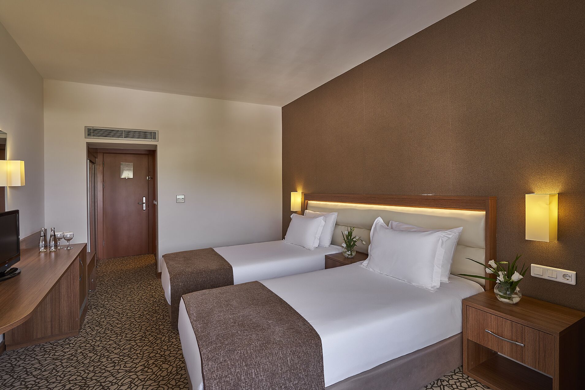 A hotel room with two beds, a desk, a TV, and a wall-mounted lamp.