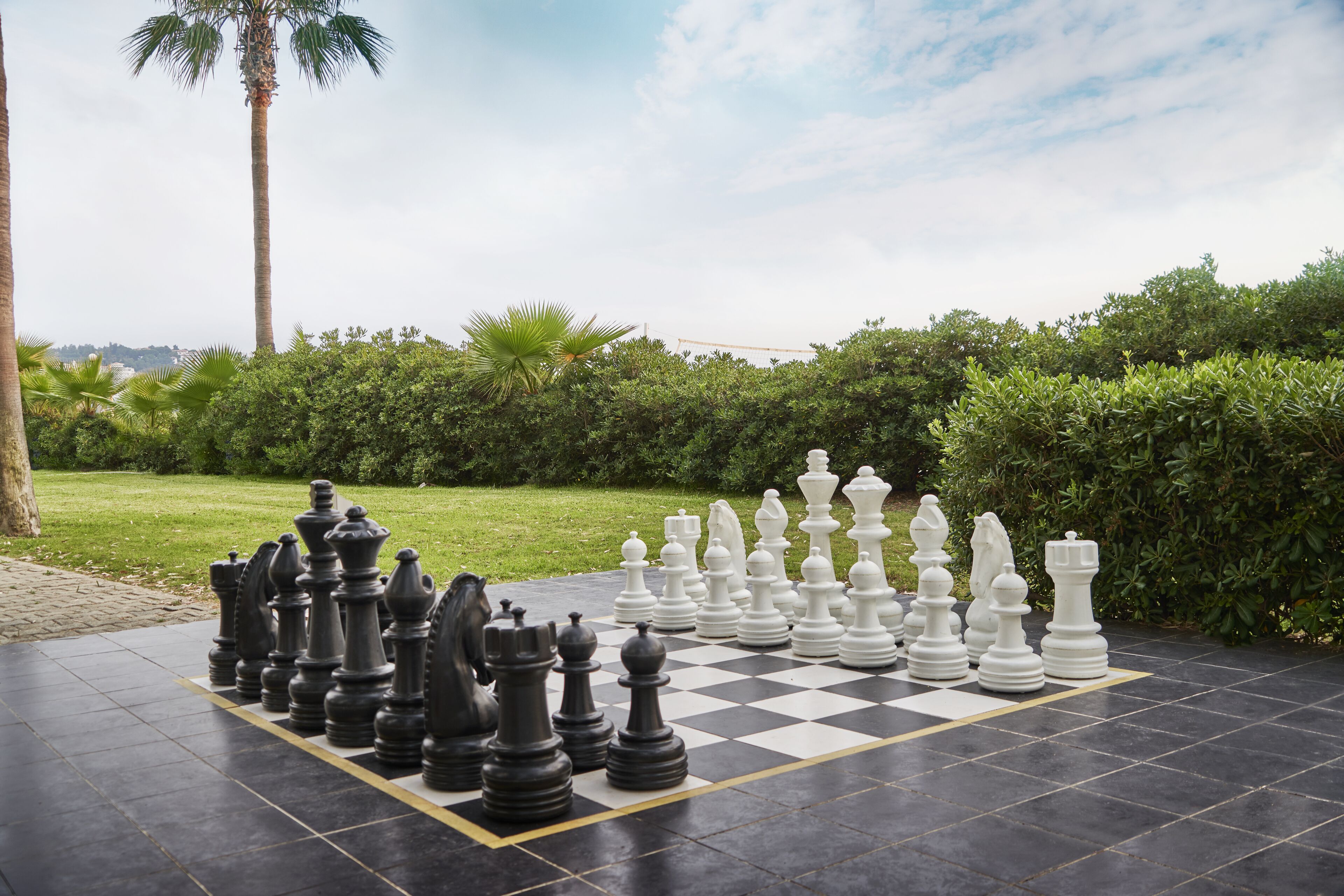 A chessboard set up outdoors with large chess pieces, surrounded by greenery and a clear sky.
