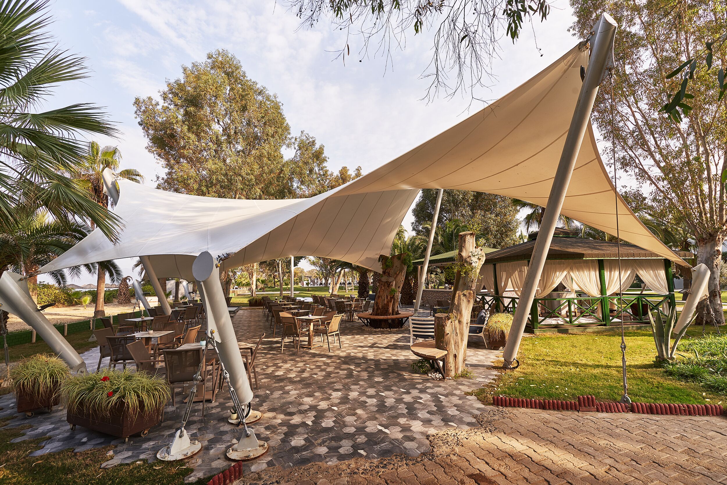 An outdoor seating area with tables and chairs under a large canopy, surrounded by greenery and trees.