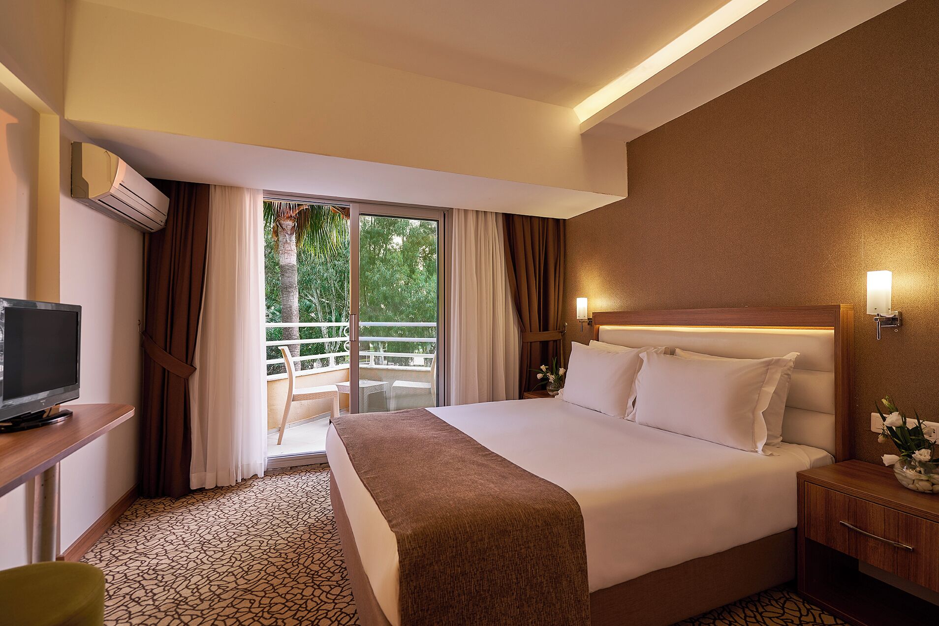 A hotel room with a bed, a TV, a desk, a nightstand, and a balcony with curtains.