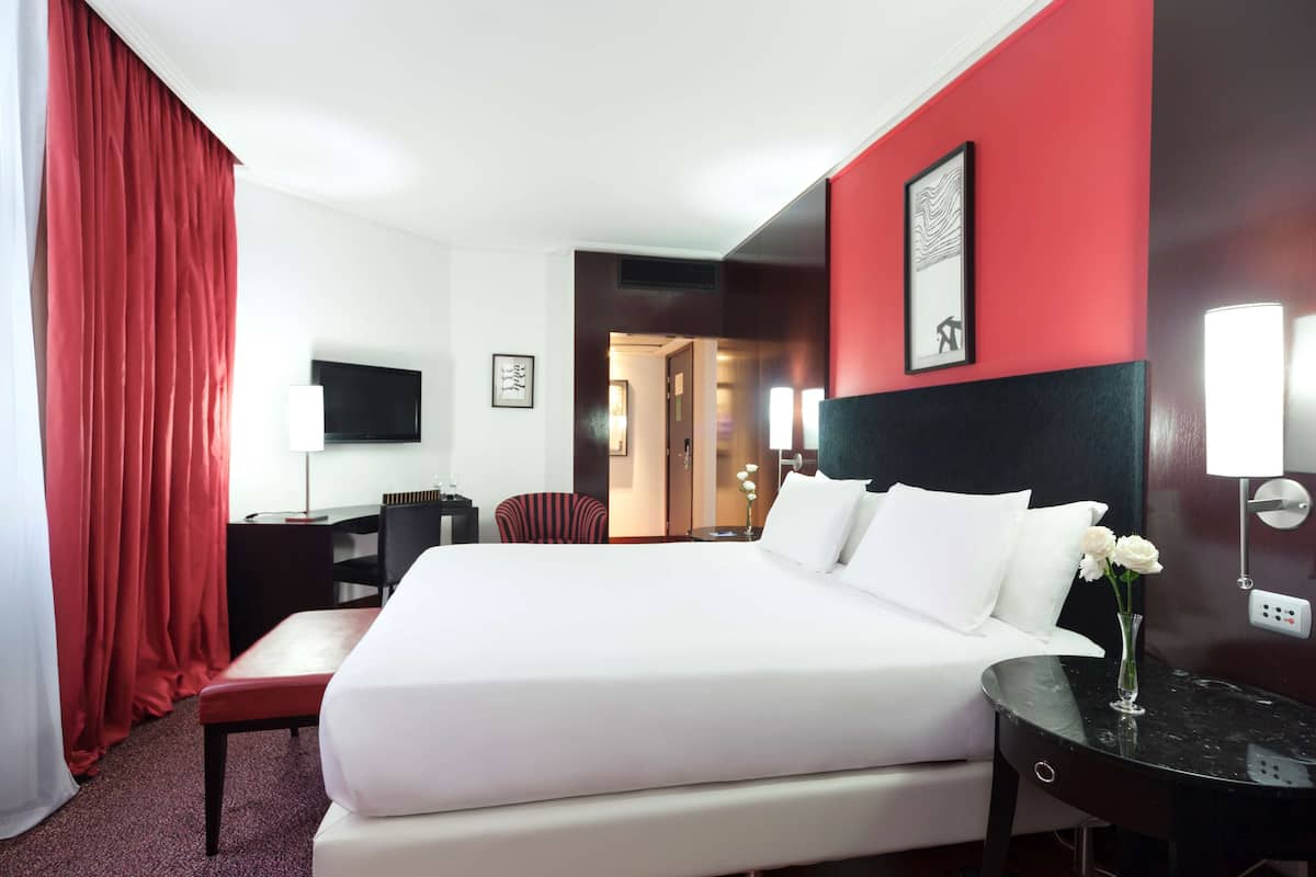 A hotel room with a large bed, a desk, a chair, a TV, and a vase with flowers.