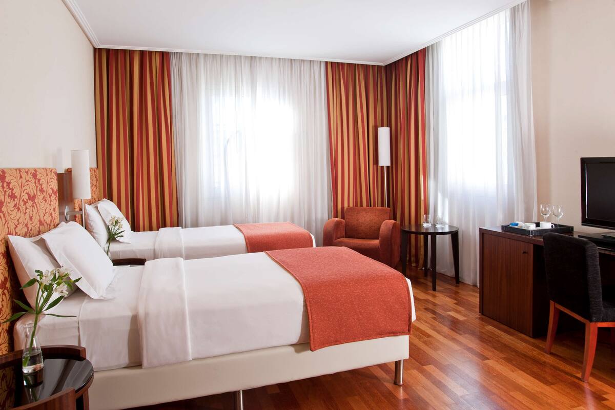 A hotel room with two beds, a desk, a chair, and a TV.