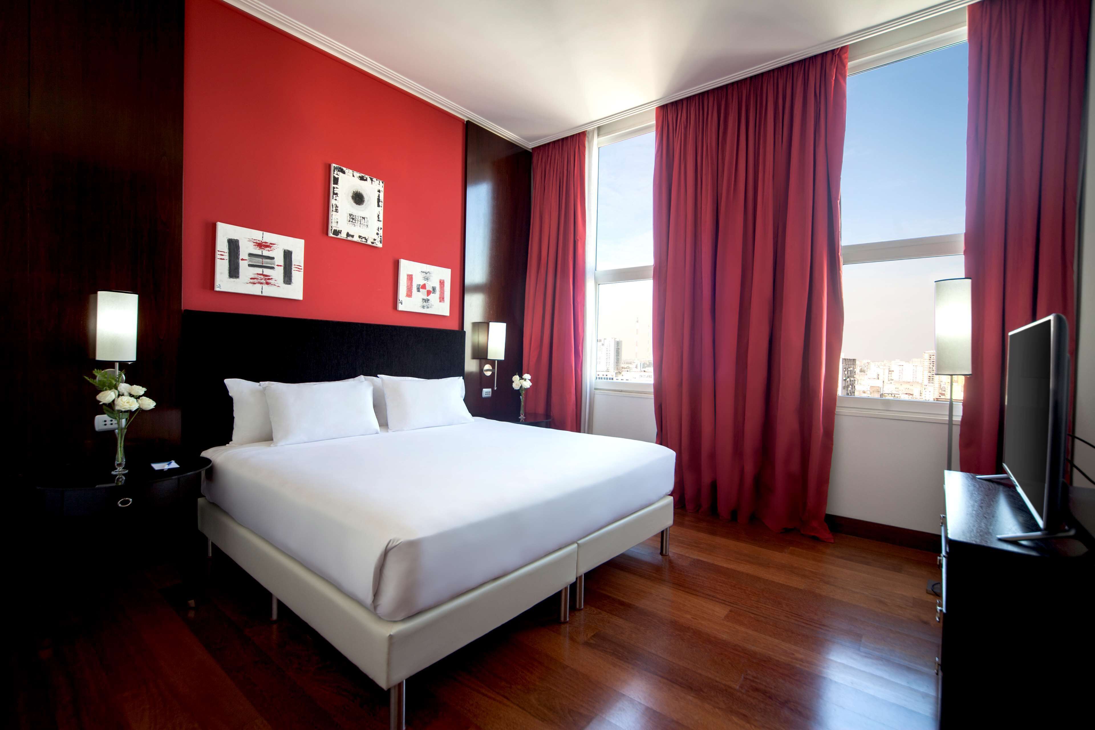 Habitación de hotel con una cama grande, paredes rojas y vistas a la ciudad.