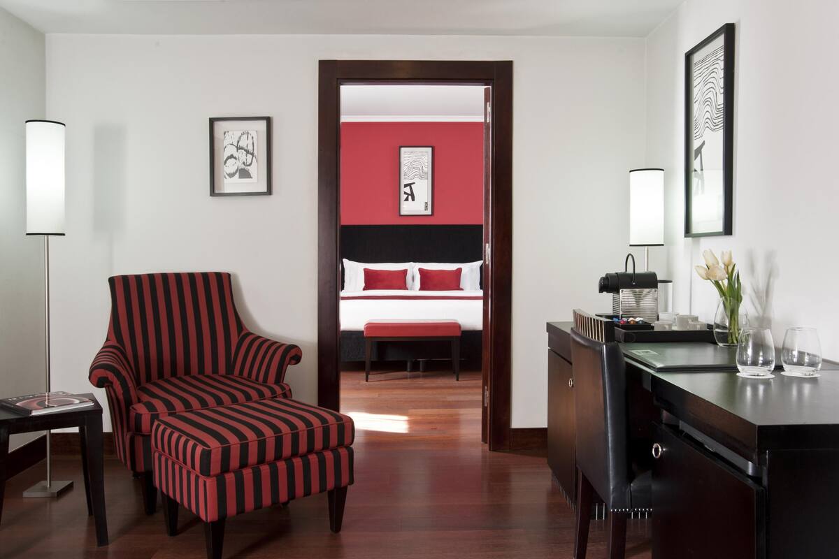 A hotel room with a striped armchair, a desk with a lamp, a bed with red bedding, and artwork on the walls.