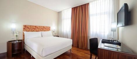A hotel room with a large bed, a desk with a chair, a TV, and a window with curtains.