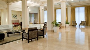 Un vestíbulo espacioso con techos altos, grandes columnas y piso pulido. Hay zonas de estar con sofás y sillones, y una gran planta en maceta en el centro.