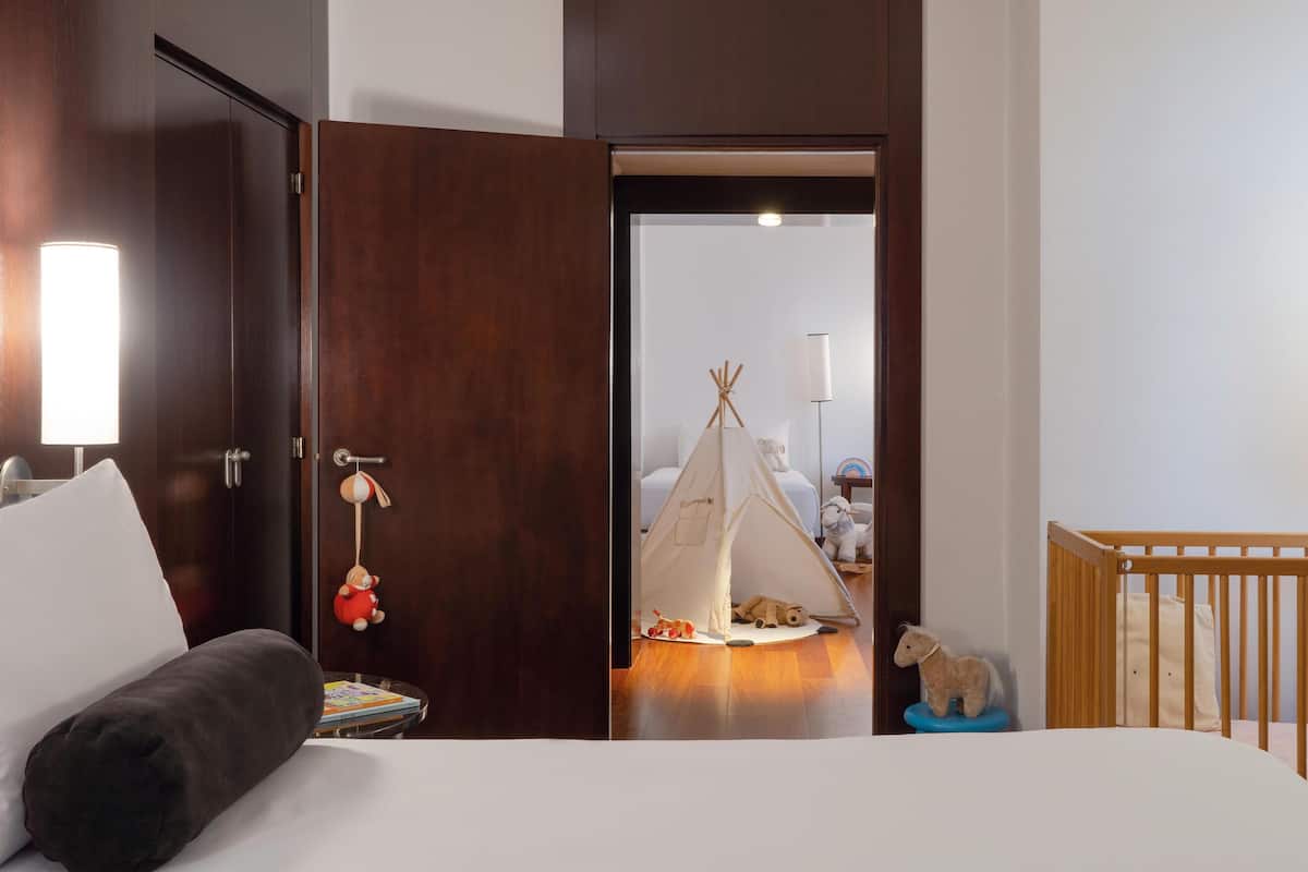 A hotel room with a bed, a nightstand, a crib, a wooden door, and a tent set up in the adjacent room.