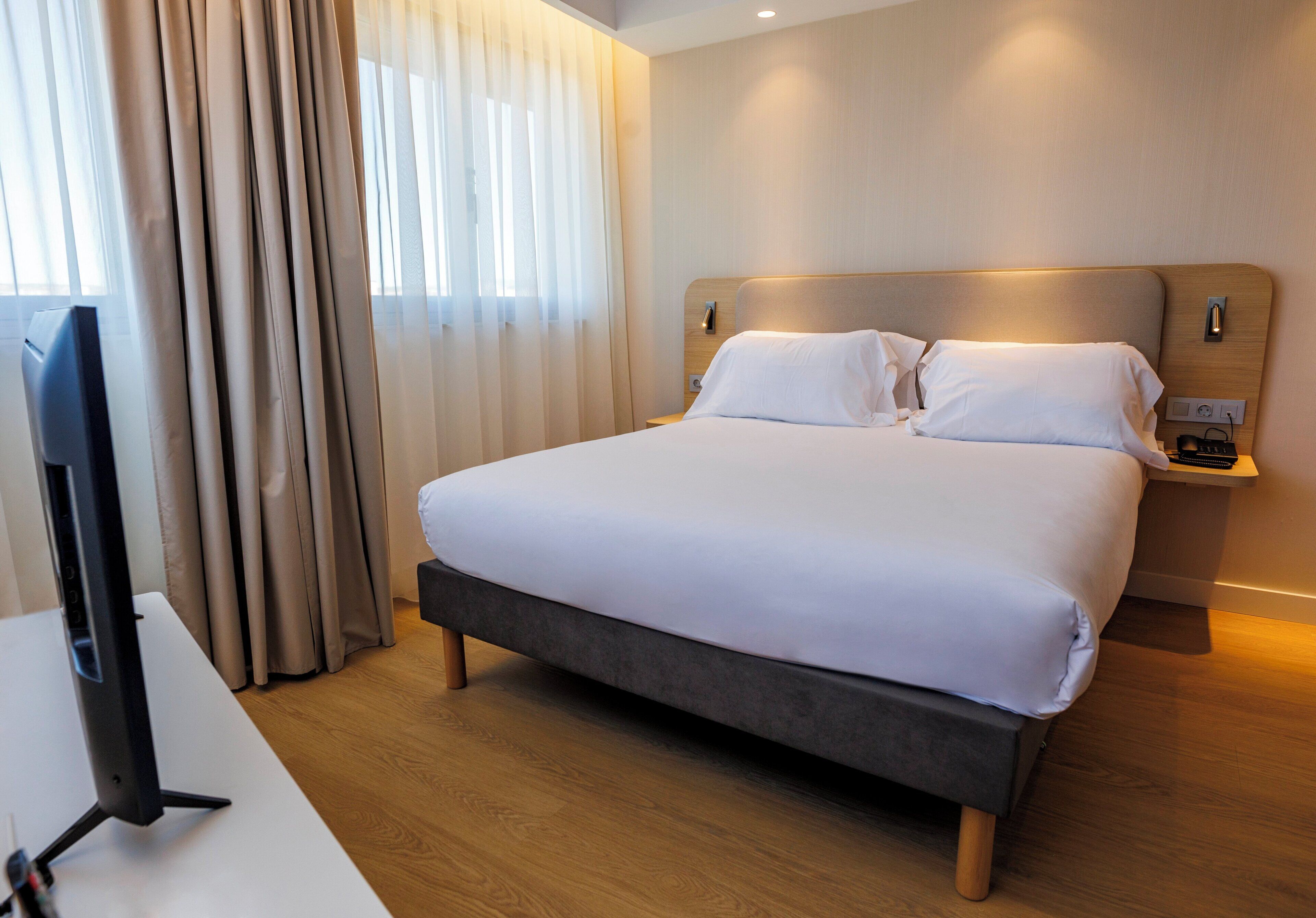 Habitación de hotel con una cama grande, una mesita de noche, un televisor y una ventana con cortinas.
