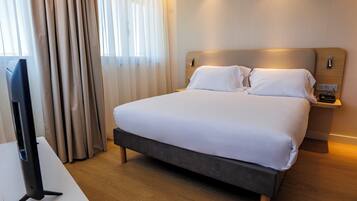 Habitación de hotel con una cama grande, una mesita de noche, un televisor y una ventana con cortinas.