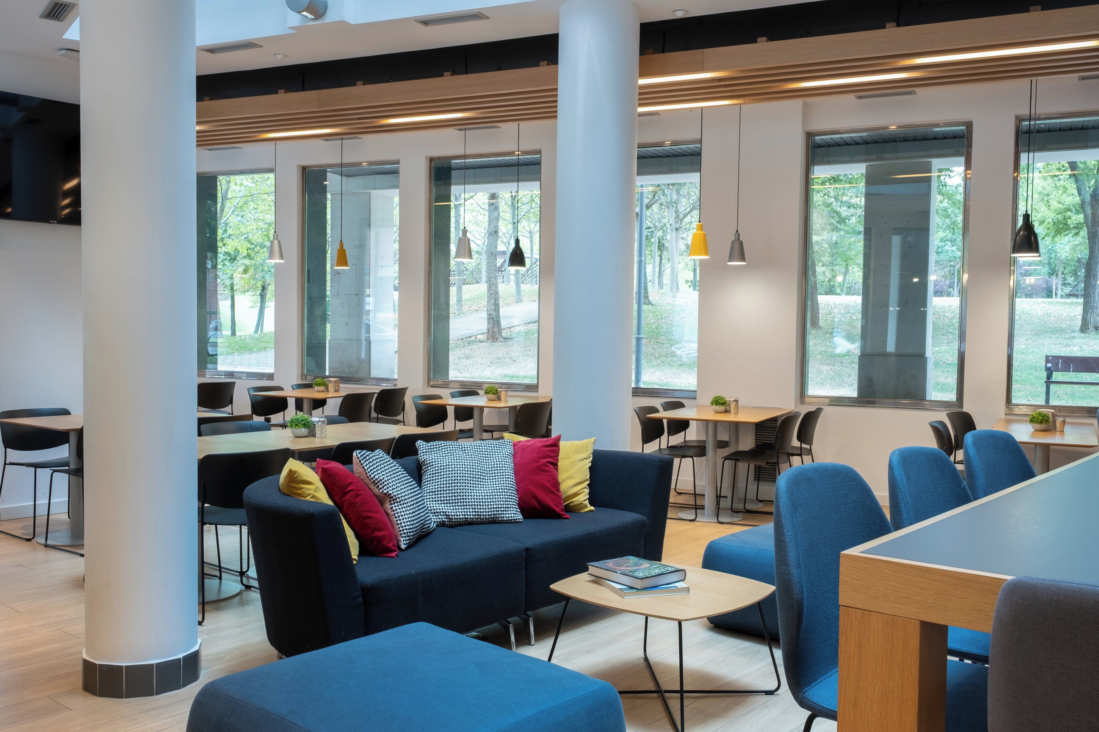 A modern lounge area with a sofa, chairs, tables, and large windows offering views of the outdoors.