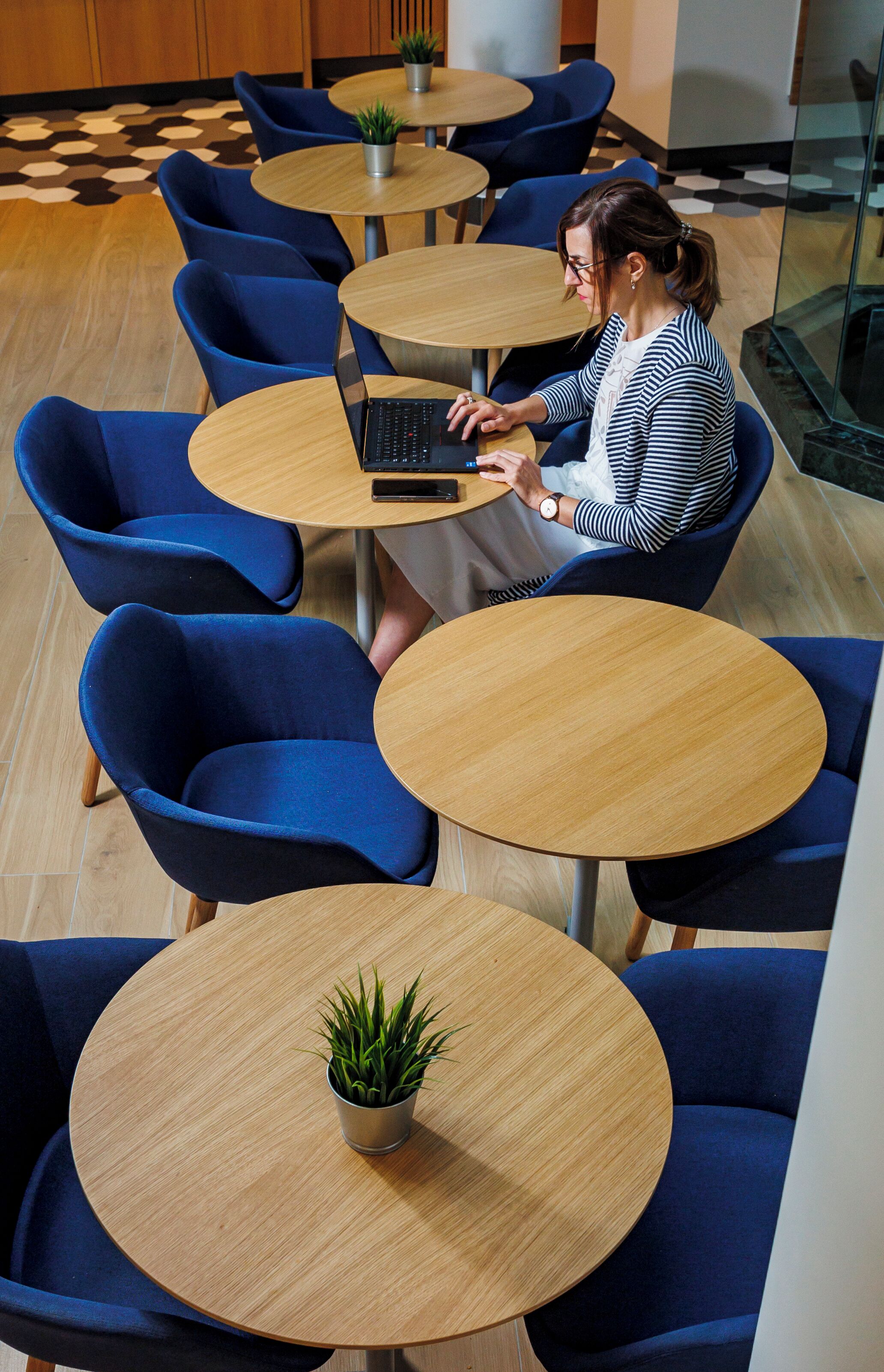 A woman seated at a round table with a laptop, surrounded by blue chairs and small potted plants.