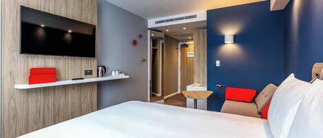 Een hotelkamer met een bed, een aan de muur bevestigde televisie, een klein tafeltje en een rode stoel.