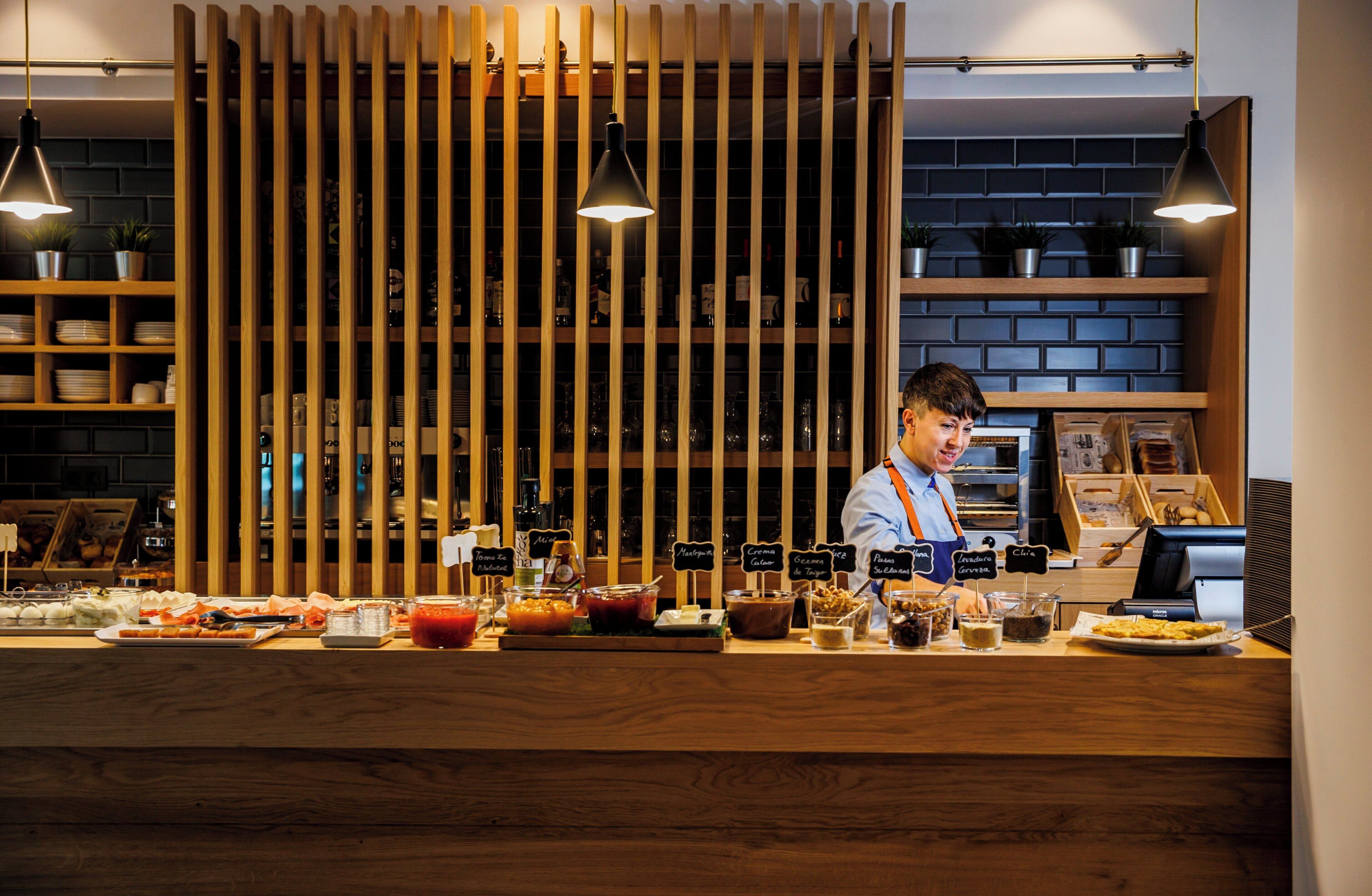 A restaurant counter with a variety of food and drink options, a cashier, and a brick wall background.