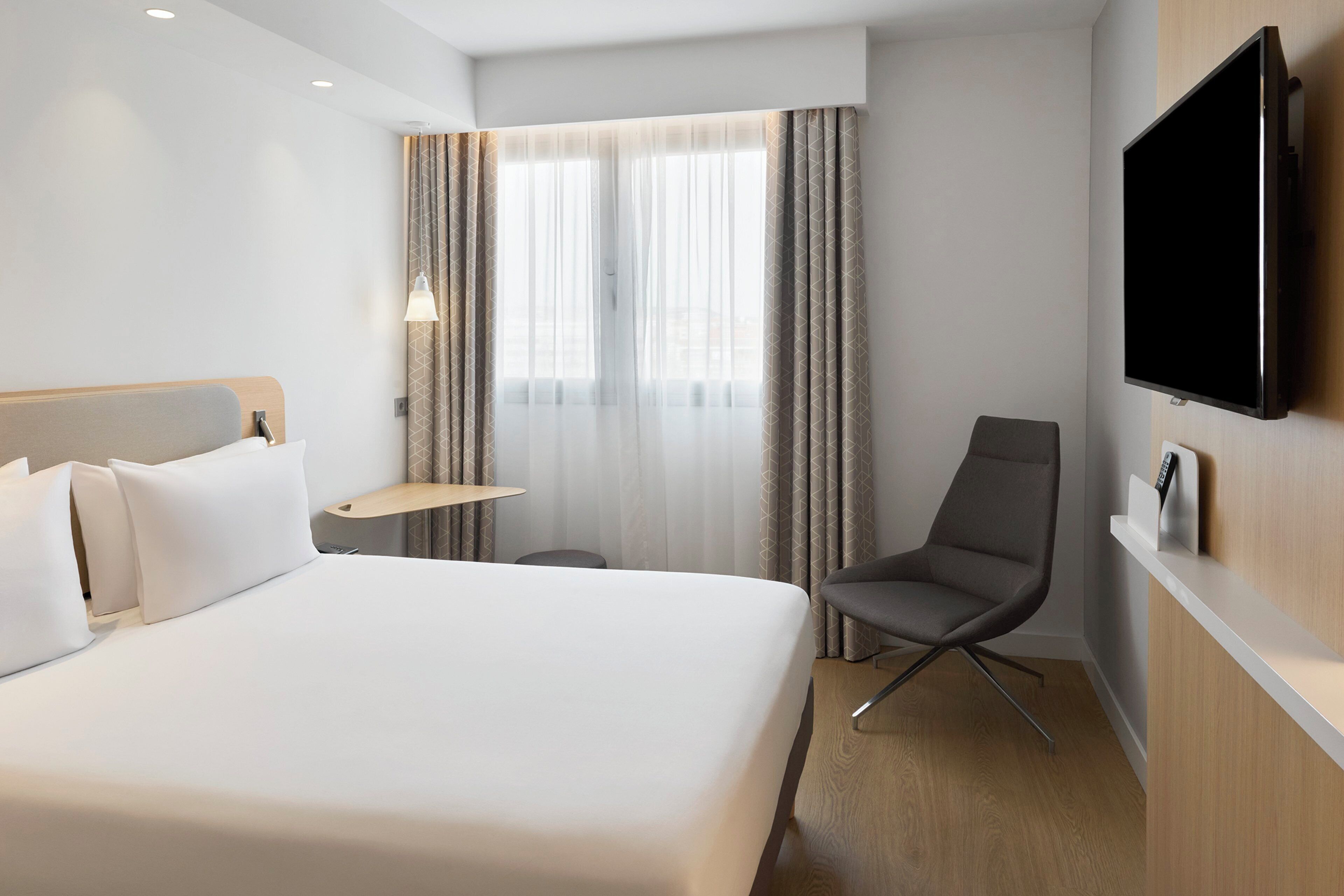 A hotel room with a bed, a desk, a chair, a TV, and a window with curtains.